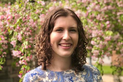 Mara Heneghan headshot pictured outside wearing a blue blouse in front of a flowering tree