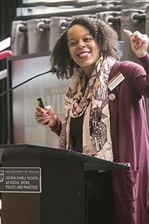 A female-presenting person smiles widely towards the camera while giving a speech at a podium.
