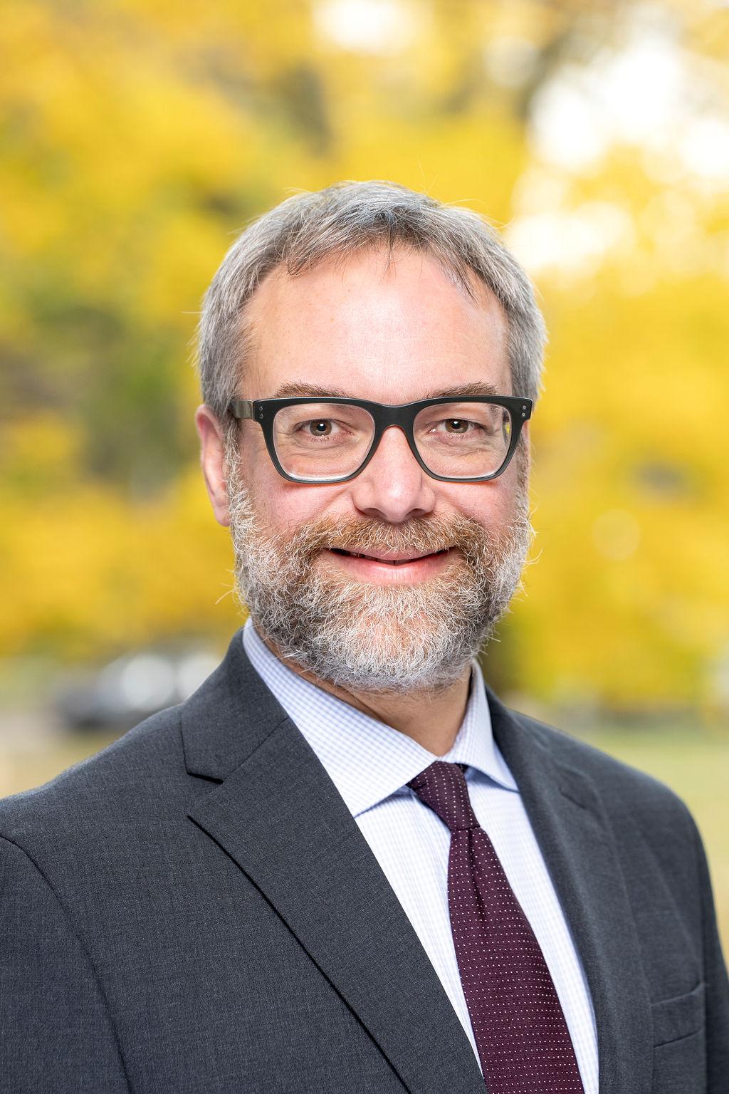 Male in grey suit with maroon tie and white shirt with glasses smiling