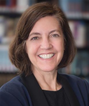 Colleen Grogan, a female-presenting person, smiles towards the camera in a library.
