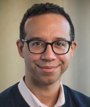 Johhny Lassalle, a male-presenting person, smiles towards the camera against a tan background.