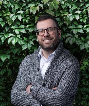 Matthew Katz in a white dress shirt and gray cardigan against a leafy background