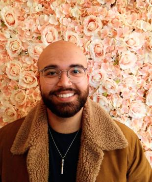 Male in brown coat with glasses smiling in front of a bed of pink roses