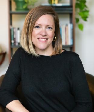 Woman sitting in front of a bookshelf with black sweater with gold hoop earrings smiling