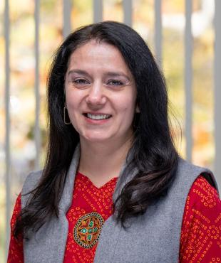 Woman in a red blouse with a grey vest with shoulder length black hair smiling in front of window with blinds